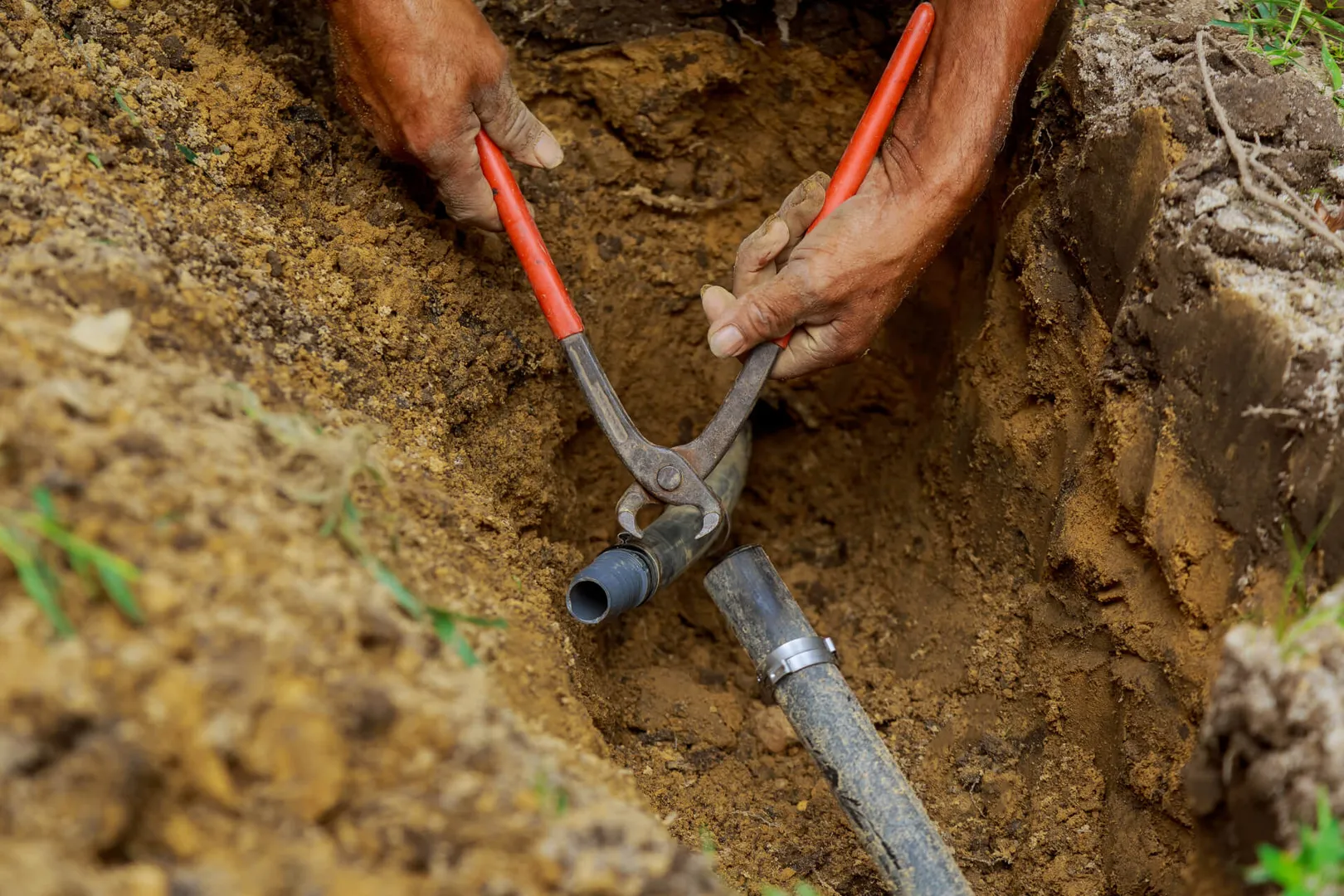 Person fixing pipe with red pliers in trench.